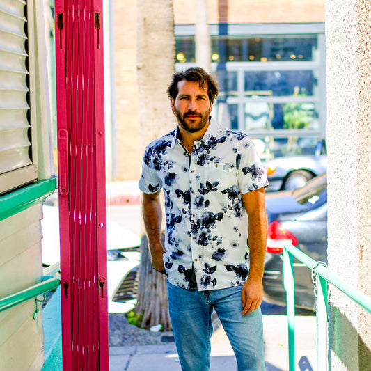 A man wearing a white short-sleeve button-down shirt with a navy blue floral pattern and light blue jeans, standing outdoors in front of a building with a bright pink door.