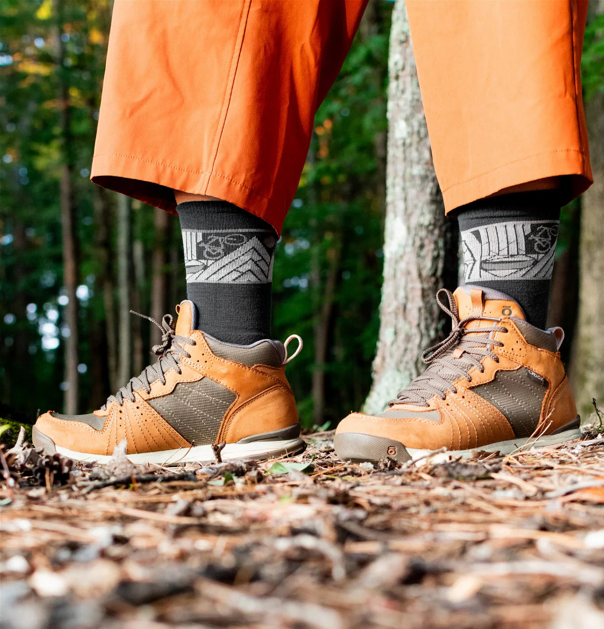 Person wearing brown hiking boots and orange pants standing on a forest floor.