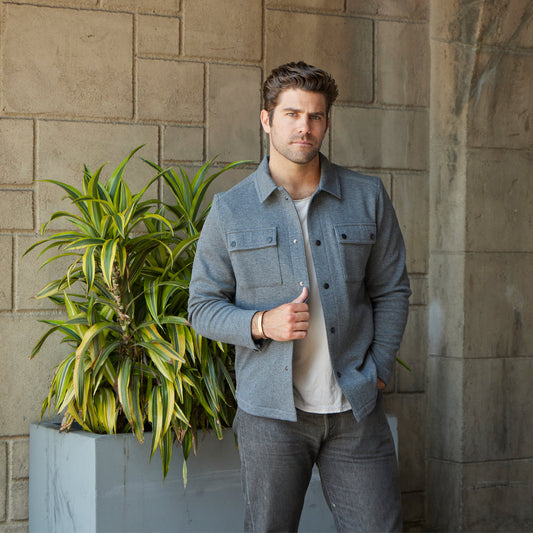 Man wearing a denim jacket standing next to a potted plant against a stone wall.