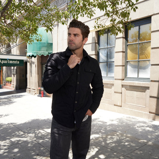 Man in a black jacket standing on a sidewalk with a building and tree in the background