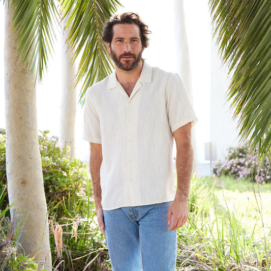 Man wearing a white shirt and blue jeans standing among palm trees and greenery.