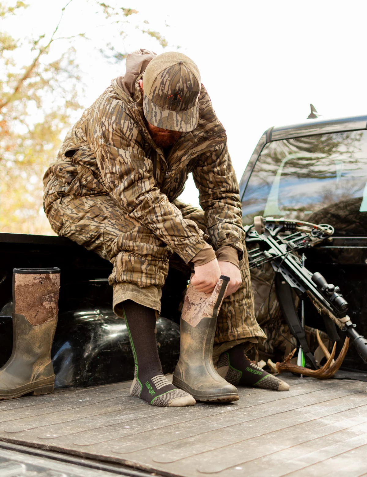 Hunter in camouflage gear putting on boots next to a truck with hunting equipment.