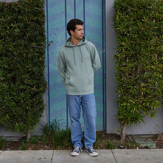 Man wearing a light green hoodie and blue jeans standing against a tiled wall with greenery.