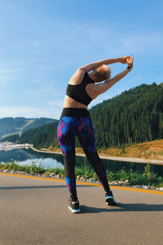 Picture of a Woman stretching with yoga pants on in front of trees and a lake.