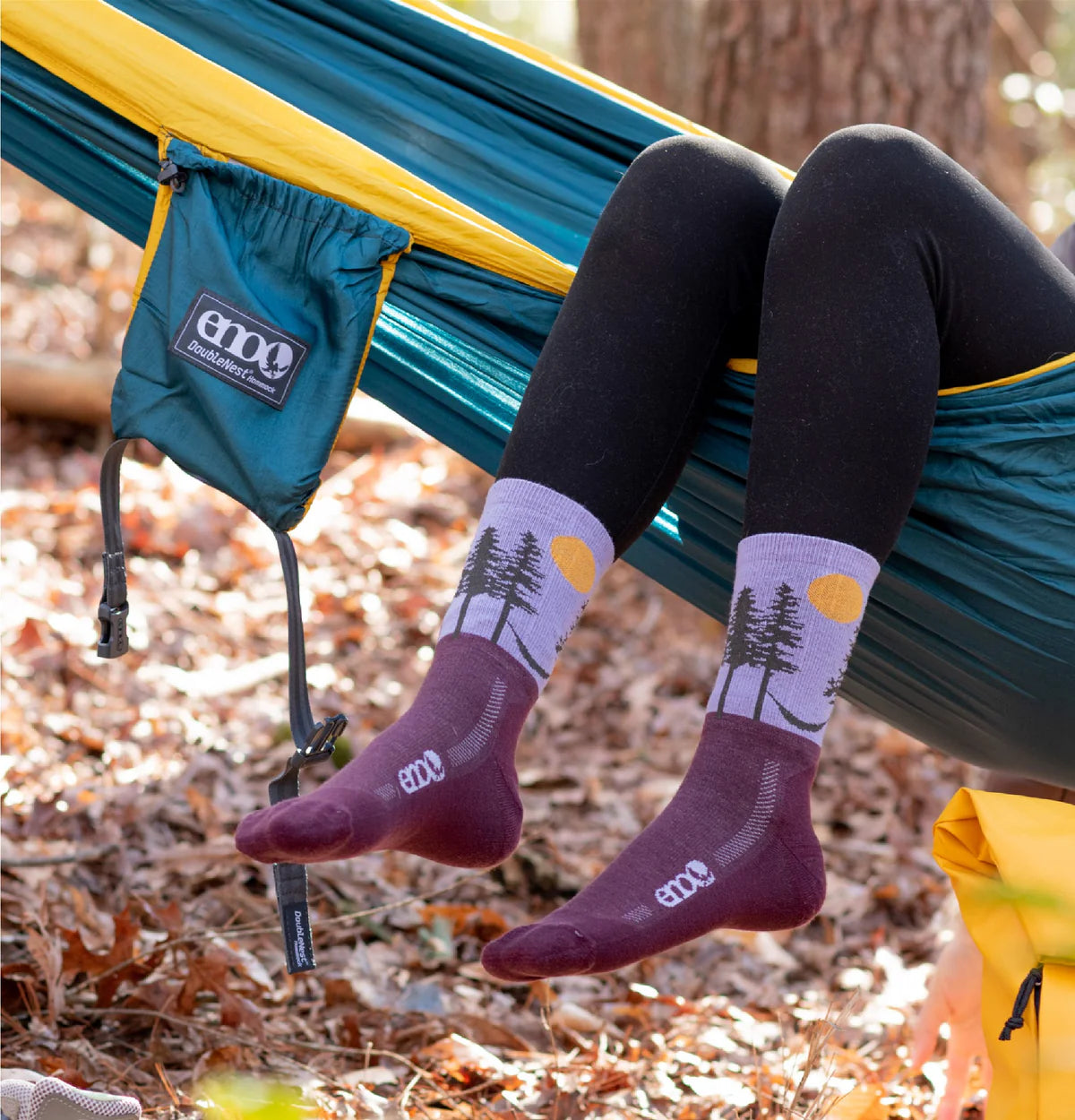 Person sitting in a hammock wearing maroon socks with tree design, surrounded by nature.