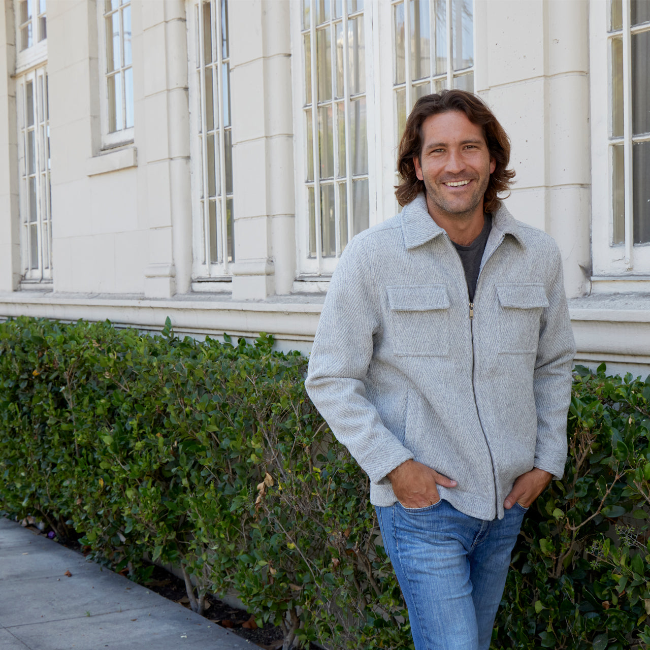 Man wearing a gray jacket and blue jeans standing in front of a building with green bushes.