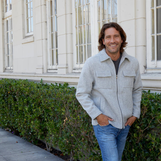 Man wearing a gray jacket and blue jeans standing in front of a building with green bushes.