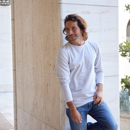 Man wearing a white long-sleeve shirt and blue jeans leaning against a stone wall.
