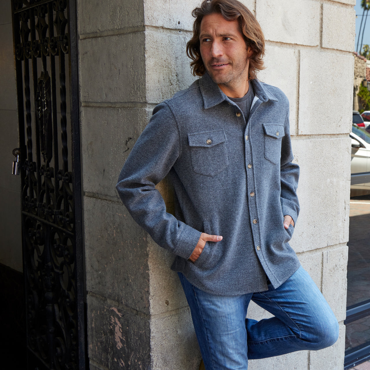 Man wearing a gray jacket and blue jeans leaning against a stone wall.