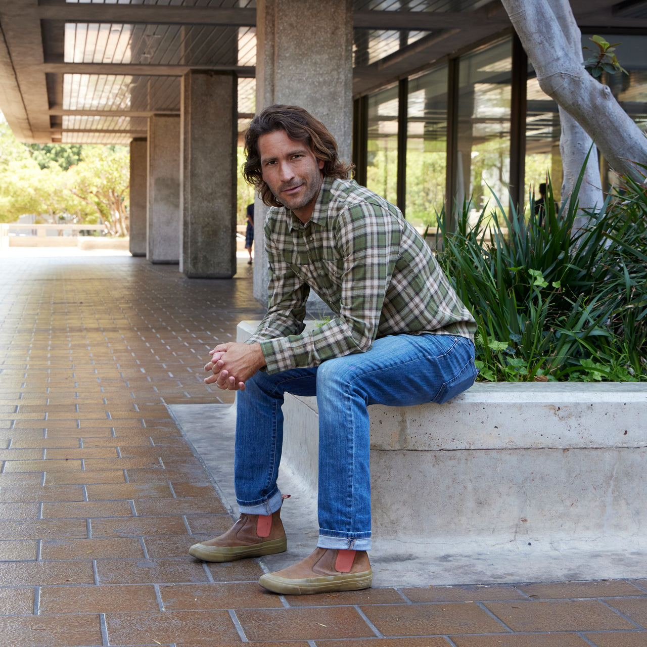 Man sitting on a concrete ledge outdoors, wearing a plaid shirt and jeans.