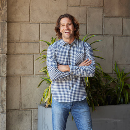 Man wearing a checkered shirt and jeans standing against a stone wall with plants.