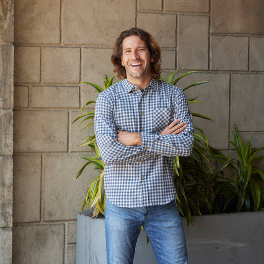 Man wearing a checkered shirt and jeans standing against a stone wall with plants.