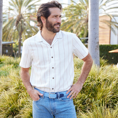 A man wearing a white short-sleeve button-up shirt with thin vertical dark pinstripes, paired with light blue jeans and a dark belt, standing outdoors in front of green plants and palm trees.