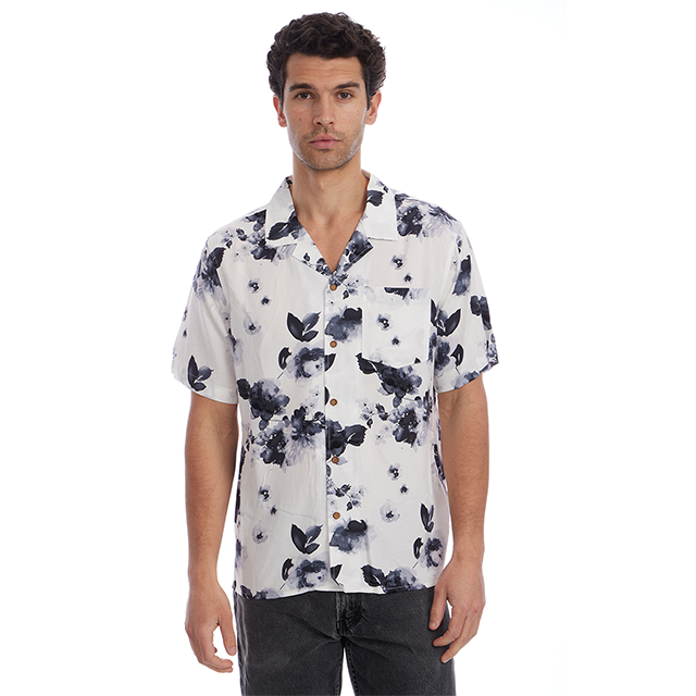 A man wearing a white short-sleeve button-up camp shirt with a navy blue watercolor floral print, paired with dark gray jeans, against a solid white background.