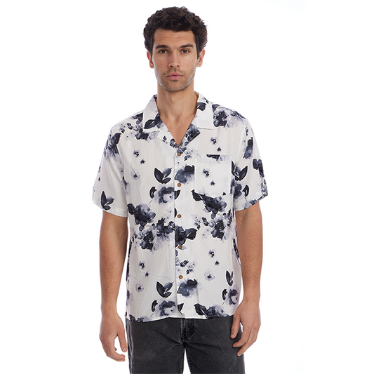 A man wearing a white short-sleeve button-up camp shirt with a navy blue watercolor floral print, paired with dark gray jeans, against a solid white background.
