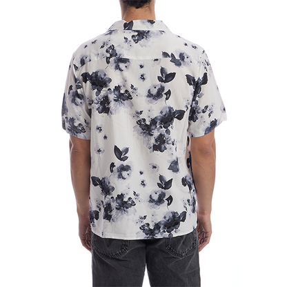 A man viewed from the back wearing a short-sleeve button-up shirt with a black and gray floral watercolor print on a white background, paired with dark gray jeans.