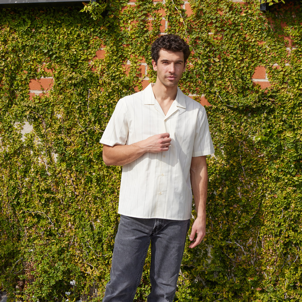 Man wearing a light-colored shirt and dark pants standing against a green wall with brick accents.