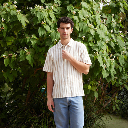 Man wearing a striped shirt and jeans standing in front of green foliage