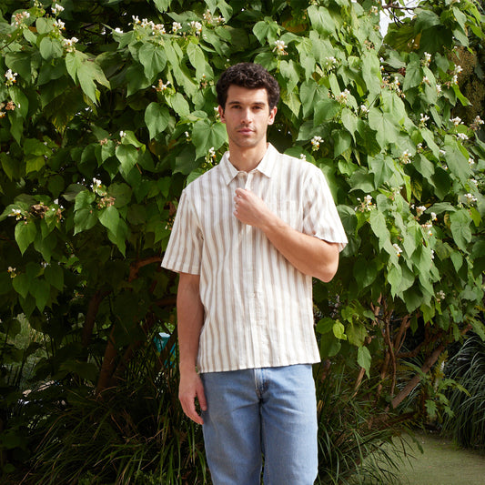 Man wearing a striped shirt and jeans standing in front of green foliage