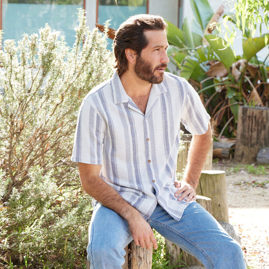 Man sitting outdoors in a garden wearing a striped shirt and jeans.