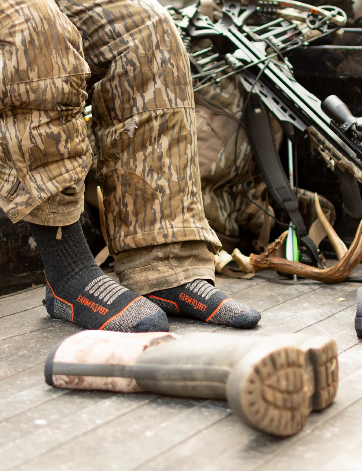 Person in camouflage gear with a rifle, wearing black socks with orange accents, on a wooden floor.