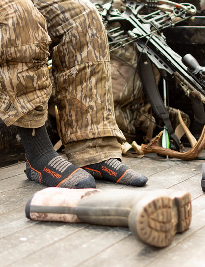 Person in camouflage gear with a rifle, wearing black socks with orange accents, on a wooden floor.