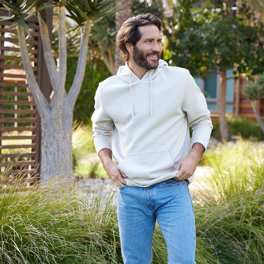 Man wearing a white hoodie and blue jeans standing outdoors with greenery around