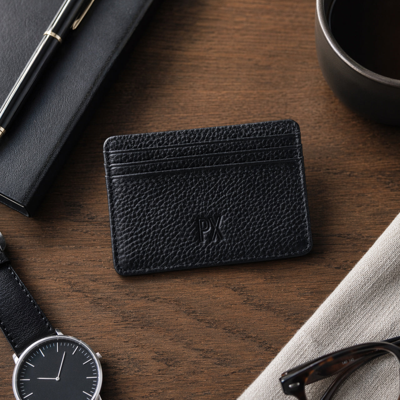 A black textured leather card holder wallet with an embossed 'PX' logo, displayed on a dark wood desk surrounded by lifestyle props including a watch, notebook, pen, and eyeglasses.