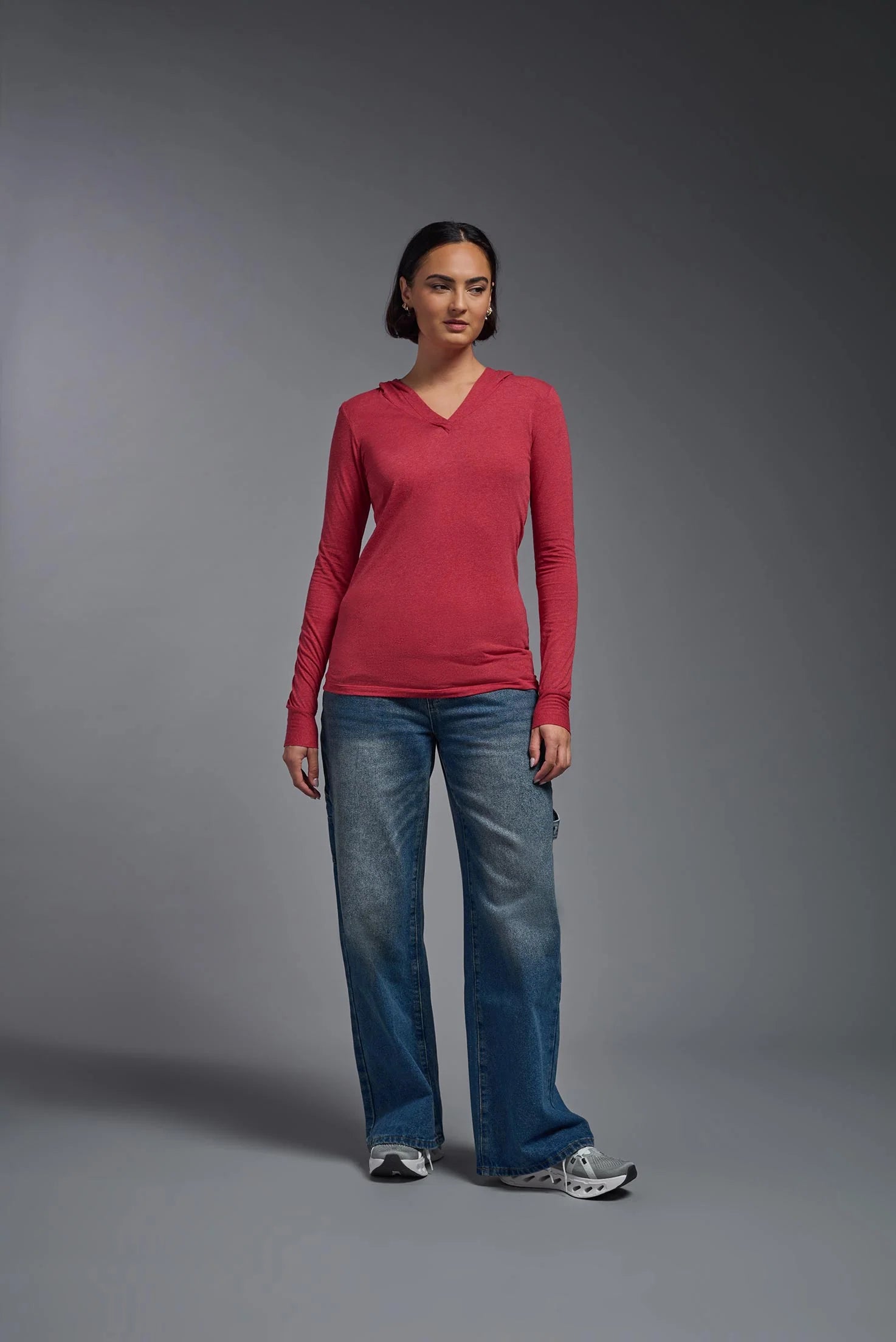 Woman wearing a red long-sleeve top and blue jeans against a gray background