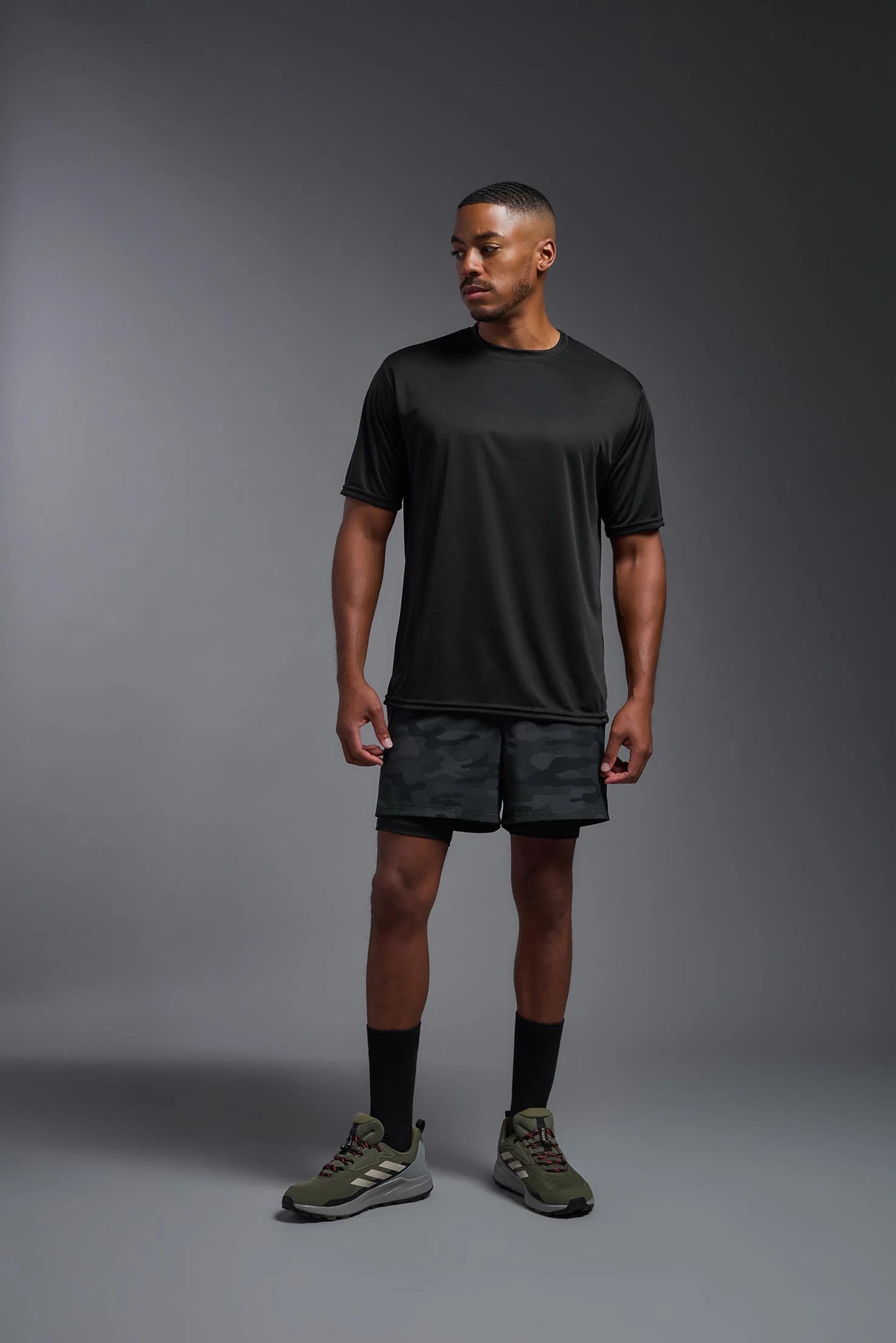 Man wearing a black t-shirt, shorts, and athletic shoes on a gray background