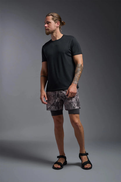 Man wearing a black t-shirt and patterned shorts standing against a gray background