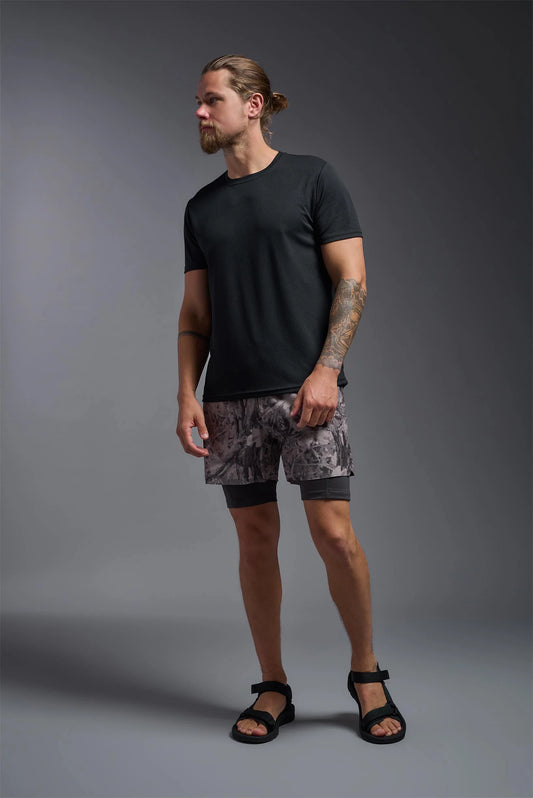 Man wearing a black t-shirt and patterned shorts standing against a gray background