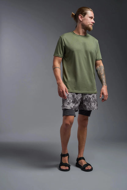 Man wearing a green t-shirt and patterned shorts standing against a gray background