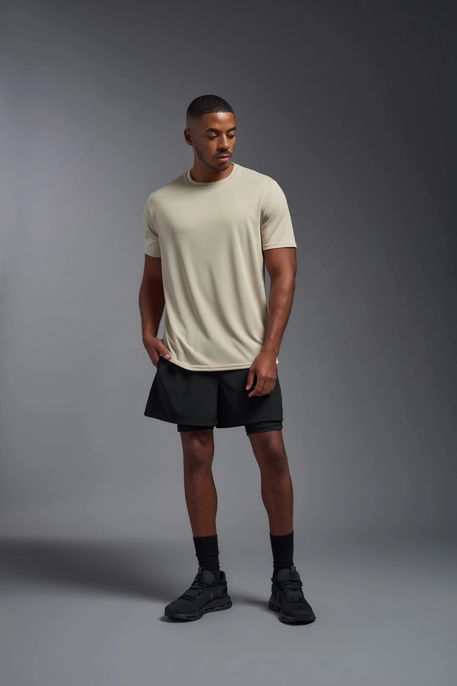 Man wearing a beige t-shirt, black shorts, and black shoes on a gray background