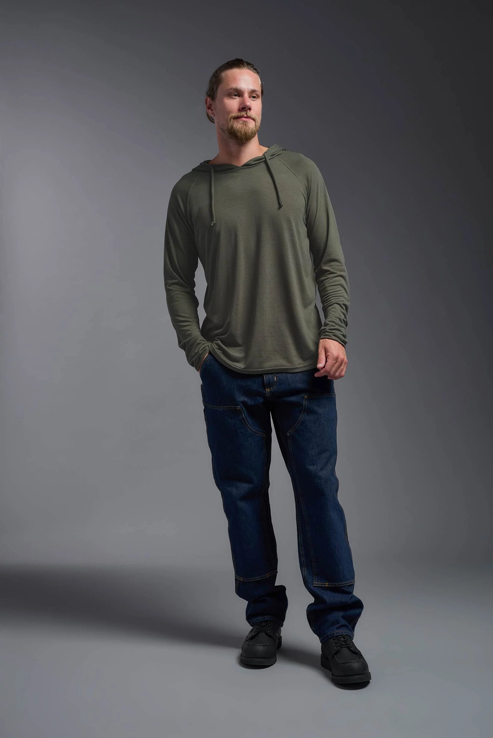 Man wearing a green hoodie and blue jeans standing against a gray background