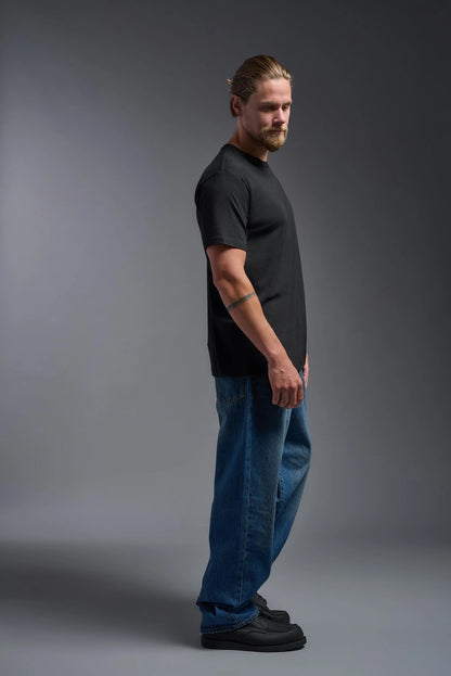 Man wearing a black t-shirt and blue jeans standing against a gray background