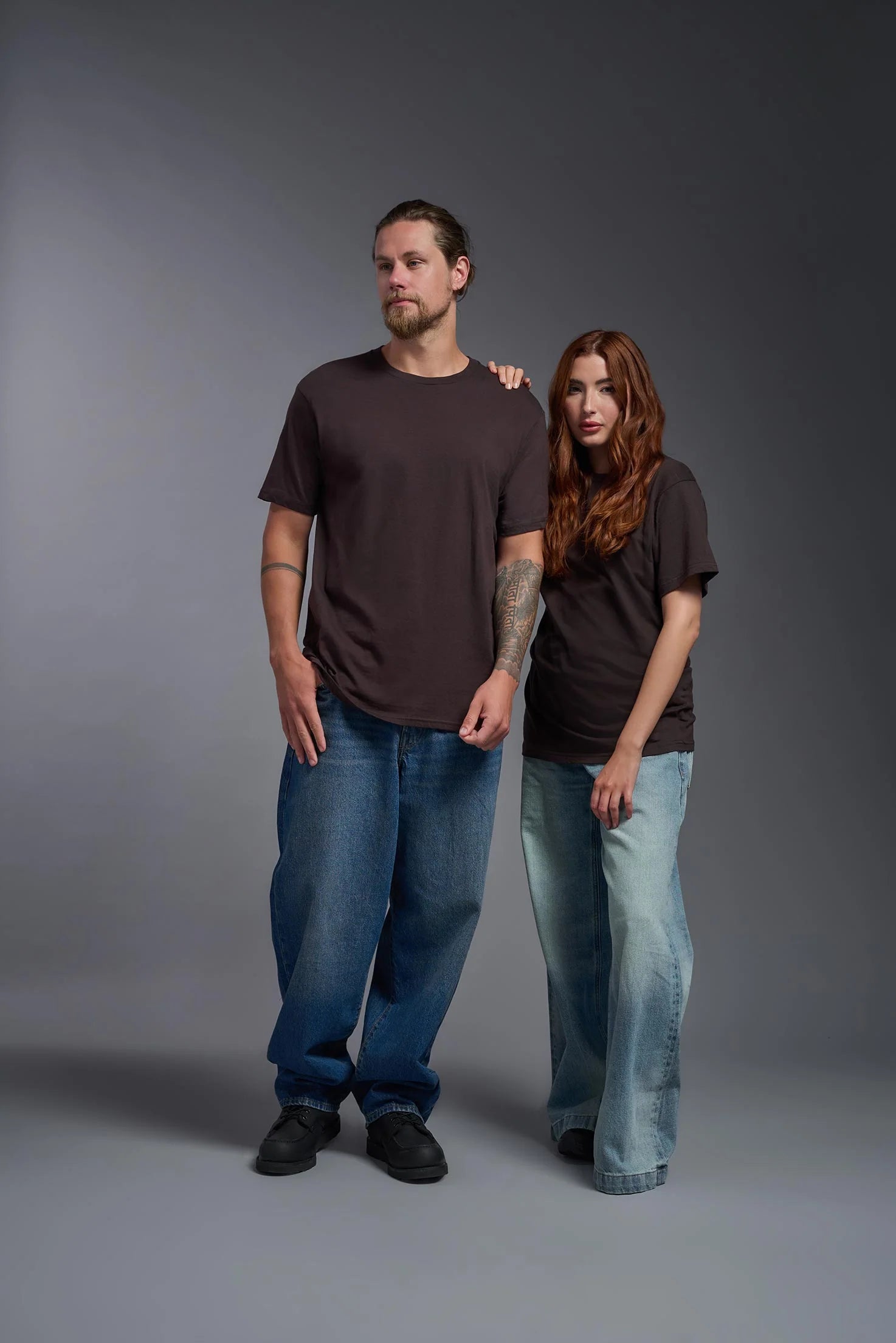 Two people wearing matching dark brown t-shirts and blue jeans against a gray background