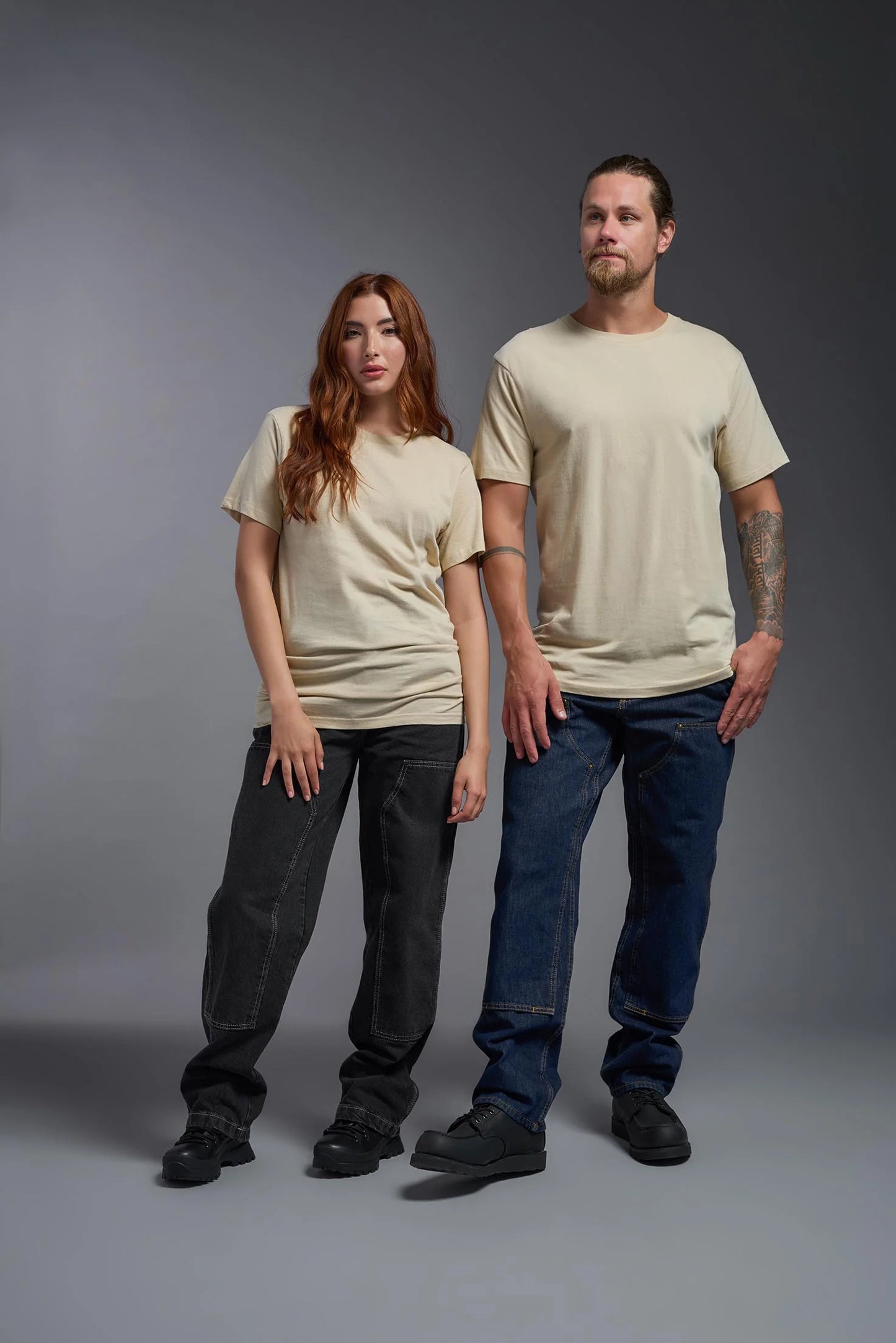 Two people wearing beige t-shirts and dark pants against a gray background