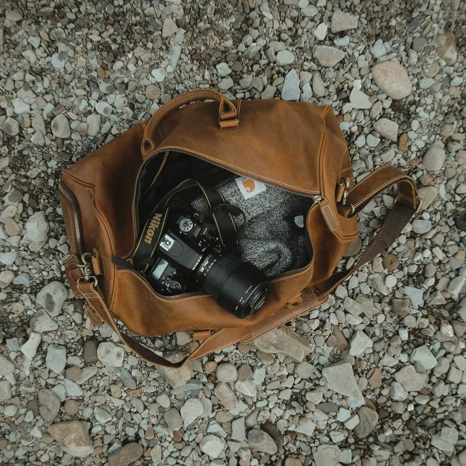 Brown leather duffle bag open to reveal camera gear inside; top handle and side straps.