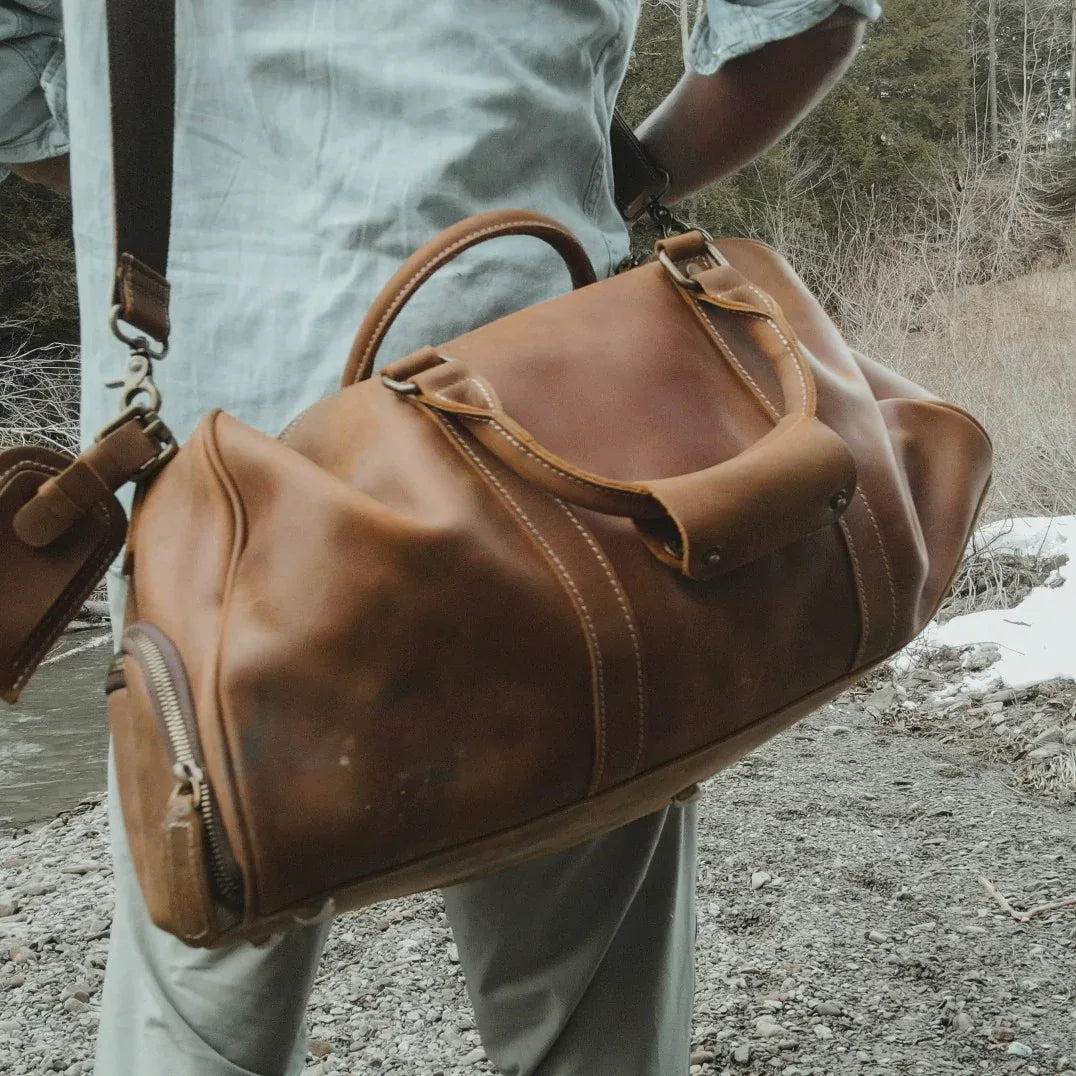 Brown leather duffle travel bag with top handle, crossbody strap, and front zipper pocket.