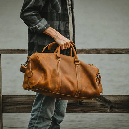Brown leather duffle travel bag with top handles, shoulder strap, and side zip pockets.