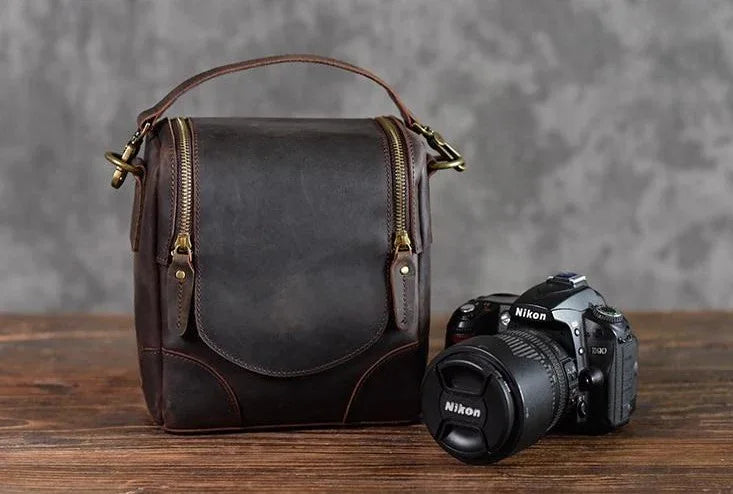 Small brown leather camera bag with curved flap and two side straps, brass hardware.