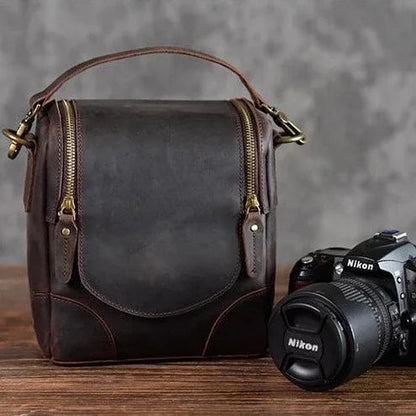 Small dark leather camera bag with front flap, zip pocket, and brass zipper pulls.
