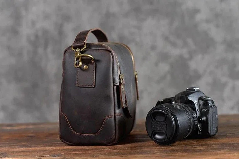 Small brown leather camera bag with single top handle, side zipper, and front label tab.