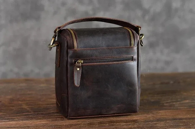 Small brown leather camera bag with front zipper pocket and top carry handle.