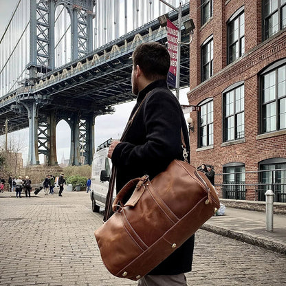 Brown leather duffle travel bag with side straps, top handles, and adjustable shoulder carry strap.