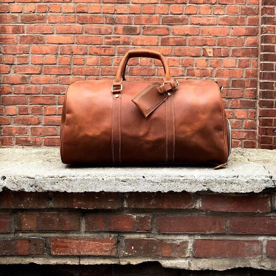 Dark brown leather duffle travel bag with top handle, front patch, and stitched side seams.
