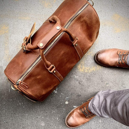 Brown leather duffle travel bag close-up showing zippers, rivets, and a long shoulder strap.