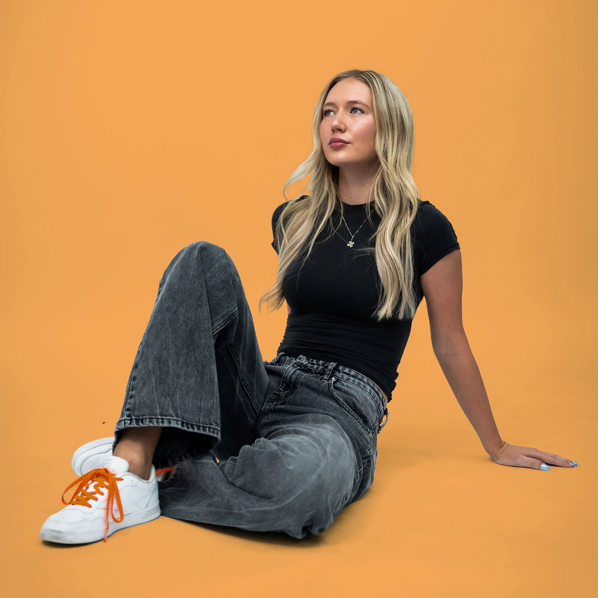 Woman sitting on an orange background wearing a black t-shirt, gray jeans, and white sneakers with orange laces.