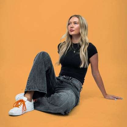 Woman sitting on an orange background wearing a black t-shirt, gray jeans, and white sneakers with orange laces.